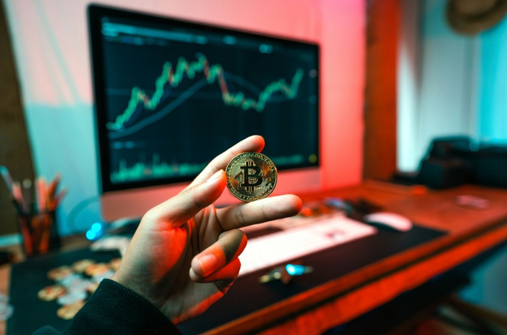 Mano sosteniendo una moneda de Bitcoin frente a un monitor con gráficos de trading